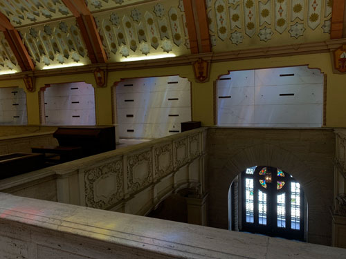 [Under an elaborately decorated
ceiling, colored light shines through recessed stained glass on walls lined with
graves along the second floor above the side aisles of a church]