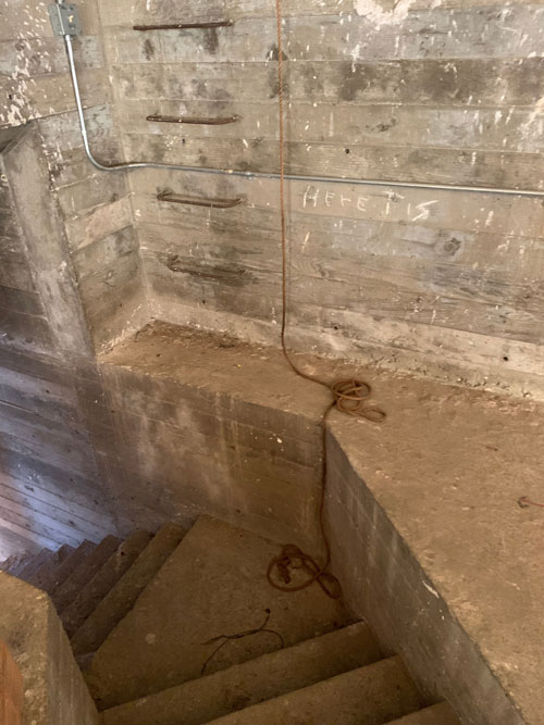 [View down a steep concrete
staircase that turns out of sight; a ladder made of rebar set into the wall
ascends from a step above the landing, and a thin rope dangles beside it from
the top of the frame. On the wall to the right in rough, angular, white letters
are the words "Here it is."]
