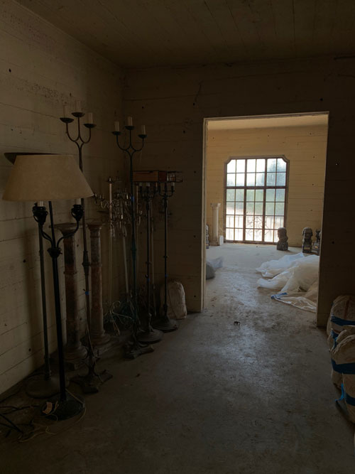 [Concrete hallway lined on one
side with plaster lumps and on the other with various kinds of standing electric
lamp, mostly in the shape of candled on a metal pole; beyond an archway, a
brighter room with a pebbled window on the floor of which is a column and
various sculptural busts.]
