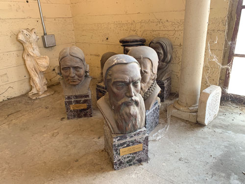 [In the center, several busts of
famous men wearing various kinds of historical costume, including
representations of John Knox and John Wesley; in the right foreground, a cracked
column connected to a rusting mirror frame by several years of tattered cobwebs;
in the left background, against the whitewashed concrete wall, a dirty marble
copy of a sculpture of a woman missing its arms and head.]