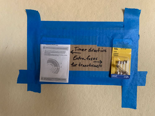 [Blue tape on a concrete wall
secures in place an instruction manual and a package of 10A fuses mounted on a
panel of corrugated cardboard on which someone has written in black marker
"Timer directions" and "Extra fuses for transformers" with arrows pointing in
the appropriate direction.]