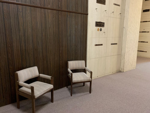 [Beige chairs on carpet before a
dark wood-textured wall beside a stone wall full of graves.]