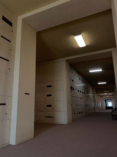 [A hallway lit by boxy light
fixtures, with the same carpet, lined with graves.]
