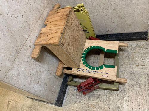 [Wooden apparatus, tucked into a
corner with a yellow "Caution: Wet Floor" sign, on which someone has written
with blue ink, transcribed below.]