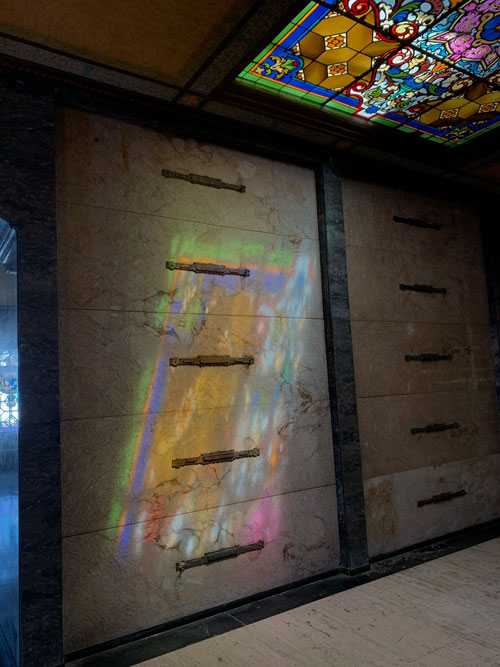 [Another view of stained glass
casting colorful light onto a wall lined with graves.]