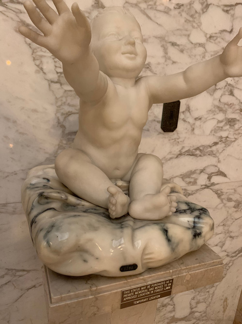 [Marble statue of an infant with
oustretched arms sitting on a marble pillow mounted on a marble pedestal against
a marble wall; on the pedestal, a sign, transcribed below.]
