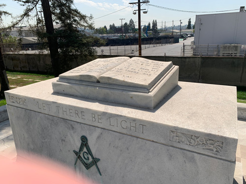 [Stone altar with sculpture of an
open book, cut with the letters "Let there be light" and a stone-and-compasses
symbol; a green lawn bordered by a barbed wire fence beyond which can be seen a
paved expanse, telephone poles, windowless industrial buildings, etc.]