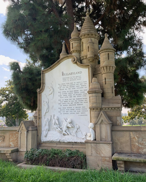 [Stone monument to "Lullabyland"
in the form of a fantastic castle with many cone-roofed turrets and a poem on a
large marble slab with relief sculpture of winged naked infants, their backs to
the viewer, ascending into the sky; at the bottom to the right a sculpture of a
naked child faces perpendicular to the sign but looks up toward it.]
