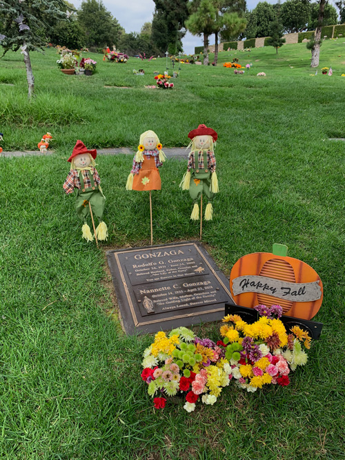 [Copper grave marker decorated
with scarecrow dolls, planters full of flowers, and a sign in the shape of a
pumpkin with the words "Happy fall"; further up the hill, dotting a plane of
green manicured grass that extends as far as one can see, are many other
decorated graves.]