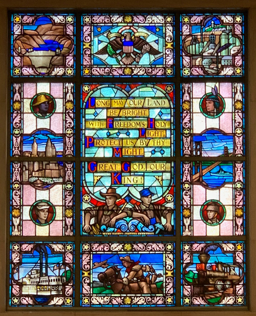 [Elaborate stained glass window
divided into several sections. In the center, the words "Long may our land be
bright with Freedoms Holy Light. Protect us by thy Might Great God our King!"
above two men facing away from each other, one dressed as a cowboy, the other as
an industrial worker. Above this, an eagle as in the seal of the United
States. Below, a man on horseback with an airplane in the background. Along the
left side, from top to bottom, an aerial view of the Hoover Dam and Lake Mead,
the head of a man in a straw hat, the Manhattan skyline as it appeared after the
construction of the Empire State and Chrysler buildings, the head of a man in a
sailors hat, and an old riverboat venting black smoke. From top to bottom along
the right side, an industrial farm with large grain silos, the head of a man
wearing a feather headdress, a ship passing beneath the Golden Gate Bridge, the
head of a man in a soldiers helmet, and an old locomotive venting black
smoke.]