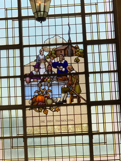 [A stained glass window. Above
the words "The First Thanksgiving," three men and a woman in Puritan dress
surround a table at which sits a long-haired figure from whose head projects two
large feathers. The table is dressed with a white cloth and on it sit a large
jug, a dish piled with colorful oblong shapes, and a dish with a cooked
turkey. In the foreground, a pile of vegetables representing the fall harvest,
including gourds and stalks of corn. In the background, a cabin with a stone
chimney from which emerges black smoke. Beyond the cabin can be seen trees and a
cloud.]