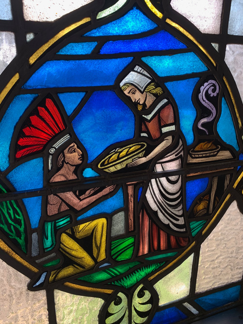 [Close-up of stained glass
window depicting a shirtless man with a large feather headdress kneeling before
and either giving or receiving a basket to or from a woman in Puritan dress; in
the background a curl of steam rises from a cooked turkey which sits in a dish
on a wooden table. Below and behind the table are some pumpkins.]