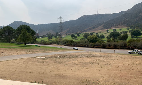 [View of a dirt expanse, a
road, overhead power lines, and low, grassy hills leading up toward the darker
chaparral of the Santa Monica Mountains, with a radio mast peeking out atop the
ridgeline.]