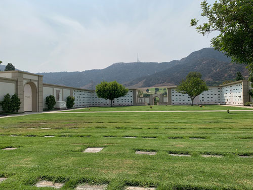 [Grassy expanse full of grave
markers within a courtyard the walls of which are also covered with grave
markers; in the distance the darker foliage of the Santa Monica Mountains and
the radio mast atop Mount Lee.]