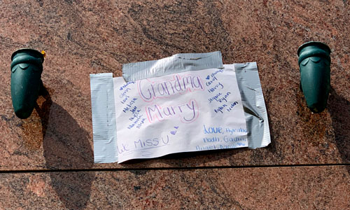 [Colorful marker on folded
white paper duct-taped to a granite slab flanked by small vases reads "Grandma
Marry, We Miss U," among other messages.]
