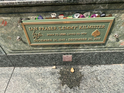 [Metal grave marker above a
stained floor commemorating "Lemmy" Kilmister, including images of his signature
and a spade as on a deck of cards. Dozens of plastic guitar picks are jammed all
around the edge.]
