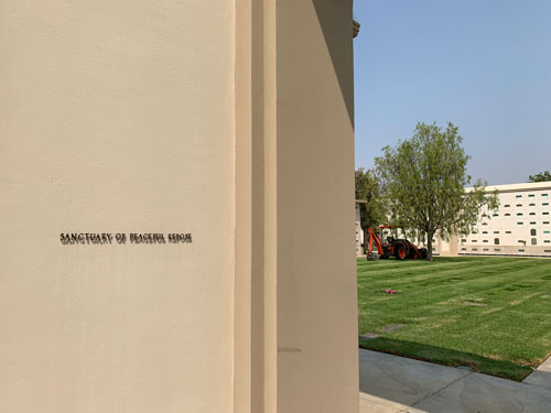 [Wall with metal lettering
announcing the "Sanctuary of Peaceful Repose" containing a courtyard with a
bright green lawn full of graves on which can be seen, under a solitary tree, a
small, orange backhoe.]
