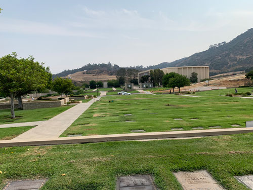 [Lawns full of copper grave
markers crossed by straight paths; in the distance, beyond a large, windowless
building, some hills of dirt leading up toward the darker chaparral of the Santa
Monica Mountains.]