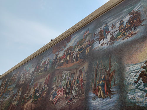 [Mosaic of various scenes, some
copied from famous paintings, representing romanticized scenes from the founding
of the United States: Flags, meetings, wartime hardship, etc., bordered at the
top with metal letters, transcribed below.]